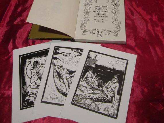 Es una edicion tapa dura entelada con tela negra y tiene un gofrado de un hacha de doble filo en el centro, vemos la portada del interior con el titulo del libro rodeado de un membrete de hojas. En la imagen tamben se observan algunas postales con xilografias que ilustran el interior del libro, unas sirenas, una persona desnuda durmiendo con gatitxs, etc...